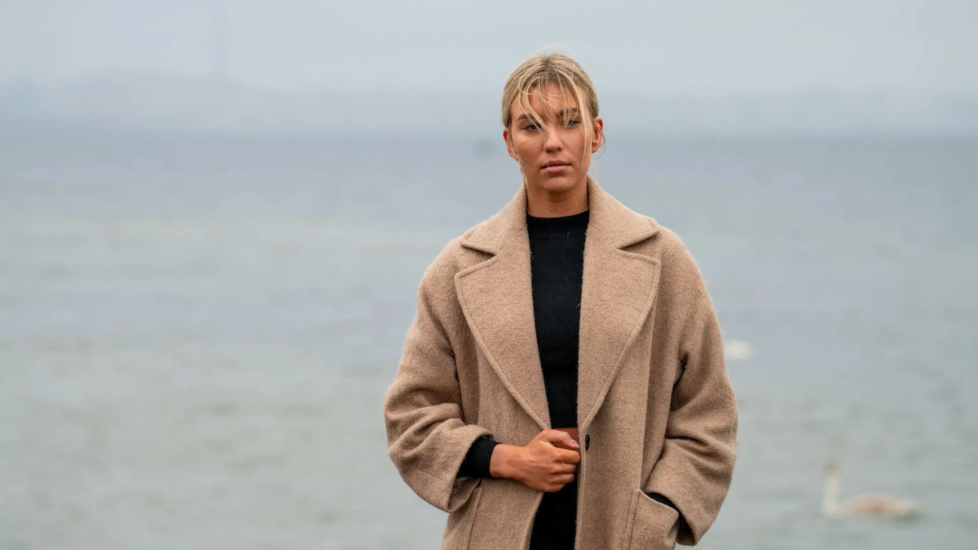 A woman in a camel wool coat stands by a misty grey sea